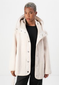 Cream-colored fleece jacket with a quilted, beige hood and snap buttons. Features a soft texture and a loose, relaxed fit.