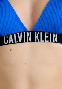 Modra prožeta bikini zgornji del z globokim V-izrezom, ki ima črn elastičen trak s tiskanjem "CALVIN KLEIN" v beli barvi.