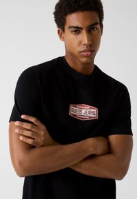 Czarny bawełniany t-shirt z naszywką z haftowanym logo "GUESS JEANS" w kolorach czerwonym i białym na środku. Krótkie rękawy i okrągły dekolt.