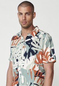 Camisa de manga corta con un patrón de hojas tropicales multicolores en verde, azul marino, naranja y crema, hecha de tejido ligero con un cierre de botones.