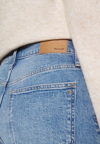 Světle modré džíny z denimového materiálu s hnědou koženou etiketou s nápisem "Madewell" na zadním pásku; mají pět kapsových provedení a viditelné švy.