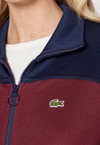 Gros plan d’une veste zippée bordeaux et marine avec un petit logo crocodile vert brodé sur la poitrine, portée sur une chemise blanche.