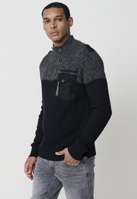 Suéter de punto acanalado en negro y gris con un cuello alto, que presenta detalles de botones y un bolsillo con cremallera en el pecho. Diseño ajustado.