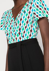 Krátký rukáv zavinovací top z hladkého materiálu s opakujícím se vlnitým vzorem v modro-zelené, bílé a hnědé barvě, doplněný o černé kalhoty s vysokým pasem.