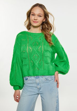 Grüner Strickpullover mit Lochmustern, Ballonärmeln und ribbeligem Saum, kombiniert mit hellblauen Jeans. Weicher Stoff und leichtes Design.