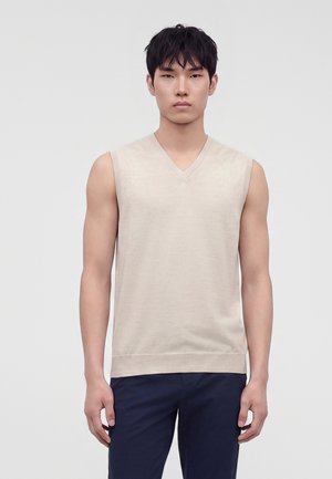 Jeune homme portant un pull beige sans manches en V et un pantalon foncé, debout devant un fond clair uni.