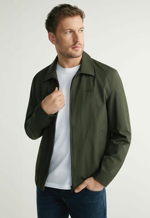 Mężczyzna w ciemnozielonej kurtce na zamek nałożonej na białą koszulę, z jedną ręką poprawiającą kołnierz, a drugą w kieszeni dżinsów, patrzący na bok.