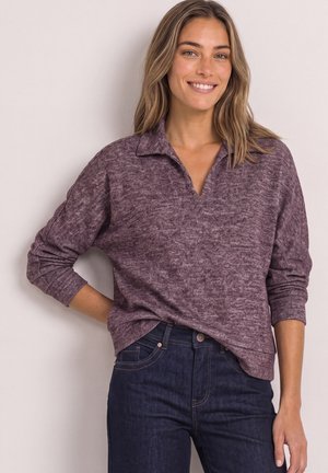 Femme souriante aux cheveux longs portant un pull violet et un jean bleu foncé, debout contre un mur clair.