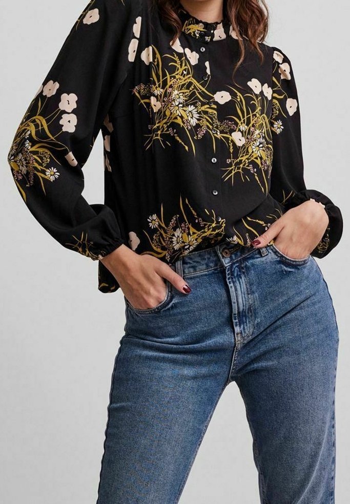 Femme portant un jean bleu taille haute et un blouse noire à motifs floraux avec des manches bouffantes et des détails de boutons, les mains dans les poches.