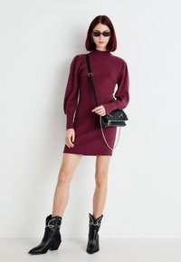 Ženska v bordo rjavi ribani obleki z napihnjenimi dolgimi rokavi, črnimi sončnimi očali, črno torbico čez ramo in črnimi vezenimi kavbojskimi škornji stoji ob belem zidu.