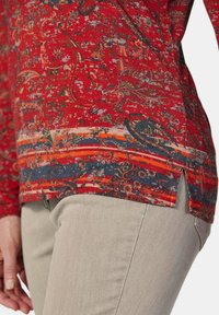 Rotes Oberteil im Paisley-Muster mit abgerundetem Saum, das Akzentstreifen in Blau und Orange aufweist. Der Stoff hat eine weiche Textur und wird mit beigen Hosen getragen.