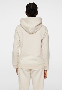 Felpa color crema con un tessuto morbido e testurizzato, dotata di un grande cappuccio e polsini a coste, abbinata a pantaloni joggers coordinati, vista da dietro.