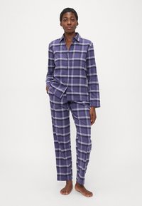 Mujer de pie descalza, vestida con un conjunto de piyama de cuadros morados de botones, con mangas largas y pantalones, con las manos relajadas a los lados.