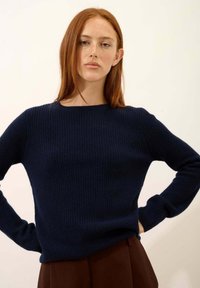 Granatowy sweter z prążkowanej tkaniny z długimi rękawami i szerokim okrągłym dekoltem, wykonany z teksturowanego materiału. Widoczne brązowe spodnie poniżej.