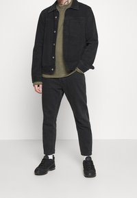 Schwarze Denimjacke mit einer Tasche, kombiniert mit einem dunkelgrauen Hemd und passenden Hosen. Schwarze Sneaker mit weißen Akzenten. Minimalistisches Design.