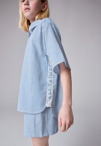 Prugasta plavo-bijela košulja kratkih rukava s ovratnikom i bočnim prorezom, s logotipom "ALLSAINTS" u plavoj boji sa strane. Uparen s prugastim kratkim hlačama.