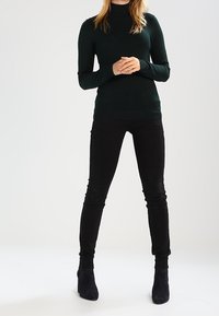 Ciemnozielony sweter z golfem i długimi rękawami, połączony z dopasowanymi czarnymi dżinsami i czarnymi botkami do kostki. Gładka faktura, minimalistyczny design.