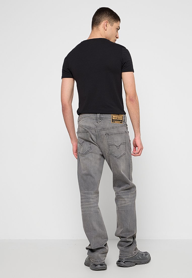 Graue Denim-Jeans mit geradem Schnitt, ausgestattet mit einem Patch auf der Rückseite und dezentem Stich auf den Taschen. Getragen mit einem schwarzen T-Shirt.