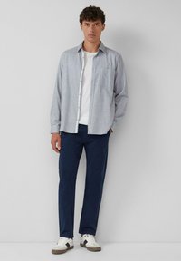 Lichtgrijze geruite overhemd met lange mouwen, wit t-shirt eronder, gecombineerd met marineblauwe broek en witte sneakers met zwarte accenten.