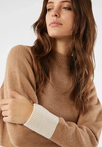 Maglione a maglia marrone chiaro con collo alto a costine e polsini a costine bianchi a contrasto. Tessuto morbido, vestibilità leggermente ampia e maniche lunghe.