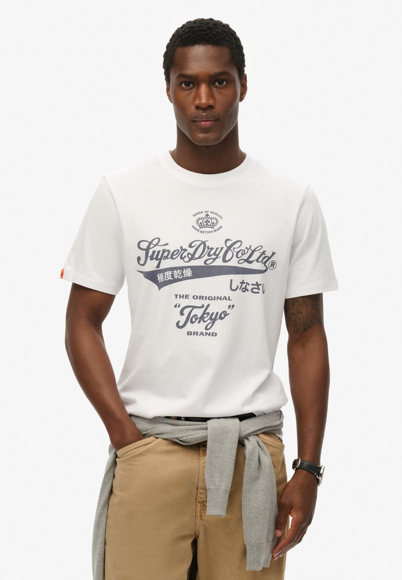 Camiseta blanca de algodón con texto gráfico en azul marino, que presenta la marca "SuperDry" y "Tokyo". Se lleva con pantalones beige y un suéter gris atado a la cintura.