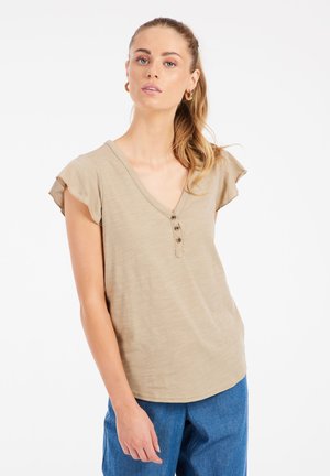 Beige Kurzarmoberteil mit V-Ausschnitt und dreiknöpfiger Leiste; weicher, leichter Stoff; sanft gerüschte Ärmelabschlüsse. Kombiniert mit blauen Hosen.