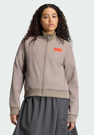 Vrouw met krullend haar die een beige jas met ritssluiting draagt met een oranje logo en een donkergrijze rok met een subtiele merkopdruk.