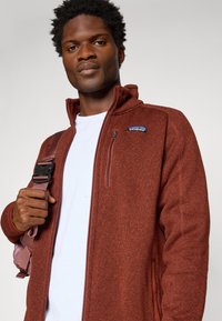 Jachetă din fleece maronie cu guler înalt, buzunar pe piept cu fermoar și patch cu logo. Modelul poartă un bretele de geantă roz deschis peste o cămașă albă.