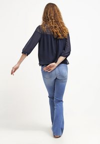 Blusa blu navy con dettagli in pizzo alle spalle, maniche a tre quarti, abbinata a jeans svasati azzurri. Capelli ricci visibili.
