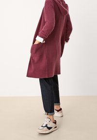 Cardigan a maglia bordeaux con cappuccio, tasche laterali e texture a coste, abbinato a pantaloni di jeans scuri e sneakers multicolori.