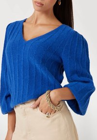Maglione blu con texture, scollo a V e design a coste. Presenta maniche voluminose. Abbinato a pantaloni beige e un bracciale dorato.