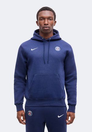 Homme portant un sweat à capuche bleu marine Paris Saint-Germain et un pantalon assorti avec des logos Nike, debout devant un fond clair uni.