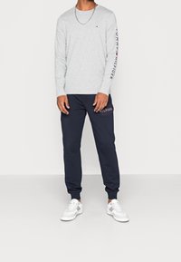 Grijs shirt met lange mouwen met een logo op de mouw, gecombineerd met marineblauwe joggingbroek met een logopatch, en witte sneakers. Eenvoudige en casual outfit.