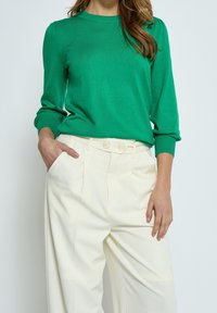 Grüner Strickpullover mit gerippten Bündchen, kombiniert mit hochgeschnittenen, cremefarbigen Hosen, die einen Doppelknopfverschluss und Falten haben.