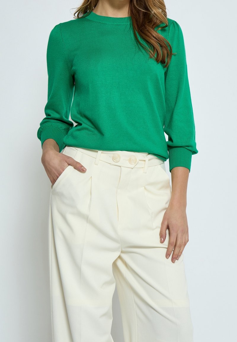Grüner Strickpullover mit gerippten Bündchen, kombiniert mit hochgeschnittenen, cremefarbigen Hosen, die einen Doppelknopfverschluss und Falten haben.