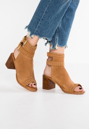 Beżowe zamszowe sandały na obcasie z otwartym noskiem, ozdobione ćwiekami, z grubym paskiem wokół kostki i drewnianym słupkowym obcasem. Widoczne są postrzępione nogawki dżinsowe.