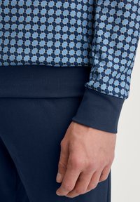Blauwe trui met een geometrisch patroon in lichte en donkere tinten blauw. Ribbelboorden in marineblauw en een zachte stofstructuur.