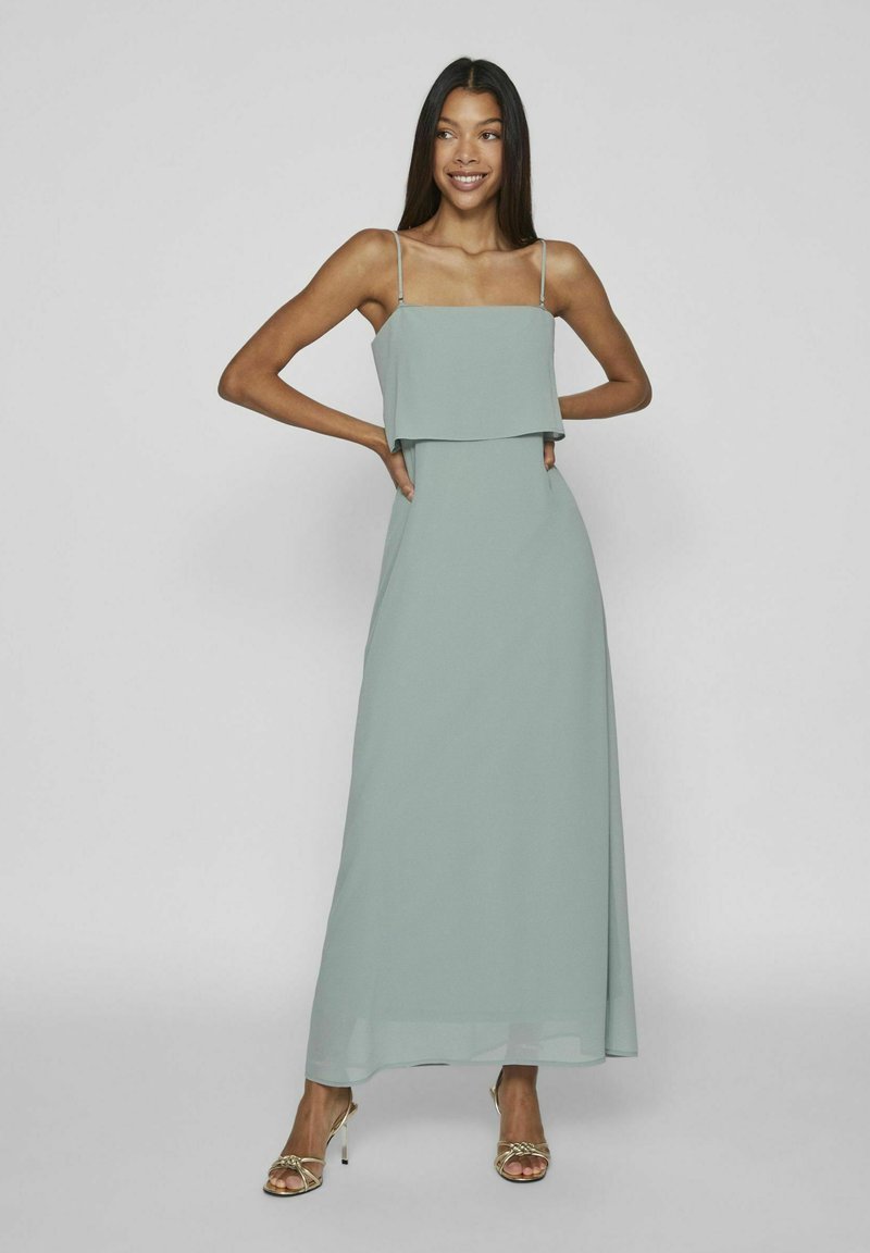 Rochie maxi verde mentă cu un top stratificat și bretele subțiri, realizată dintr-un material ușor și fluid. Accesorizată cu sandale metalice cu curele.