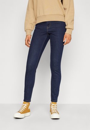 Skinny jeans in dark blue denim with contrasting stitching, paired with tan high-top sneakers with white soles. Simple sweatshirt above.
