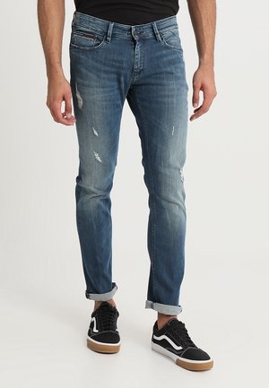 Mann trägt blaue Slim-Fit-Jeans mit hochgekrempelten Säumen und schwarze Low-Top-Sneaker mit weißen Akzenten und Gummisohlen.