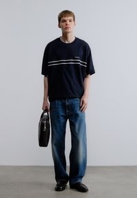 Μπλε σκούρο μπλουζάκι με κοντά μανίκια και λευκές οριζόντιες ρίγες, συνδυασμένο με ανοιχτό μπλε τζιν και μαύρα παπούτσια, κρατώντας μια μαύρη τσάντα.