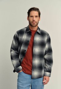 Camicia di flanella a quadri nera, grigia e bianca con bottoni frontali. T-shirt rossa a collo rotondo sotto. Jeans blu. Tessuto testurizzato.