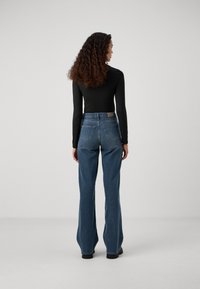 Mulher com cabelo longo e encaracolado, vestindo uma blusa preta de mangas longas e jeans azuis de cintura alta, de pé de frente para um fundo cinza claro liso.