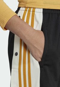 Hand i fickan på svartvita byxor med gula ränder, ihop med en långärmad skjorta i senapsgul färg på armen.