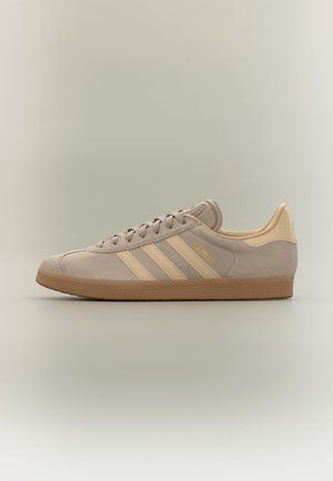 Baskets Adidas Gazelle beige avec bandes crème et semelle en caoutchouc marron, vues de côté sur un fond neutre.