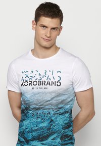 White short-sleeve t-shirt featuring a blue and black graphic design resembling water textures with text "KOROBRAND BE IN THE NOW."