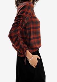 Donna che indossa un top a maniche lunghe a quadri arancione e nero con maniche arricciate e pantaloni neri, con una mano in tasca e un bracciale d'argento al polso.