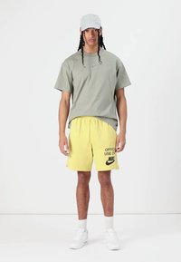 Lichtgrijze t-shirt met een klein Nike-logo, gecombineerd met gele shorts met de tekst "ENKEL VOOR OFFICIËLE GEBRUIK", gedragen met een lichte pet en witte sneakers.