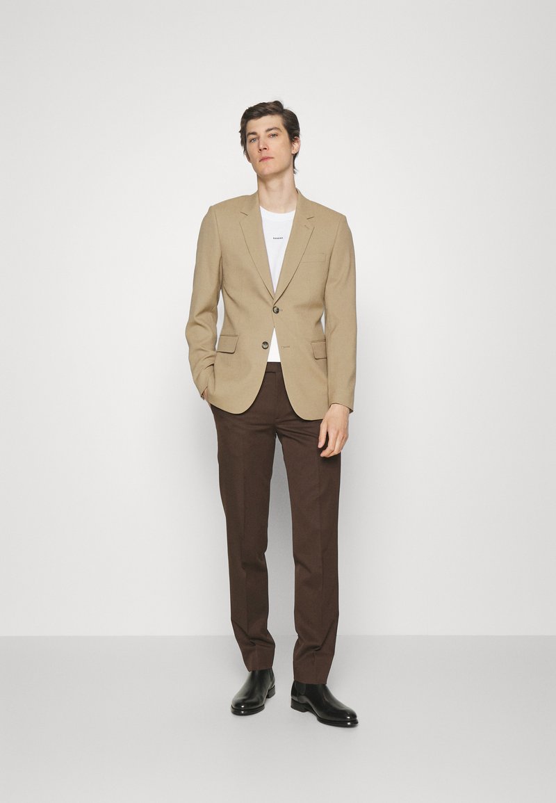 Beige blazer with a single button, paired with a white t-shirt and brown trousers. Black leather footwear completes the look.