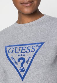 Osoba w szarej koszuli z niebieskim trójkowym logo Guess, które zawiera znak zapytania oraz napis "U.S.A. Washed Jeans" na piersi.